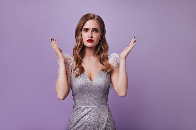 Free photo woman in silver dress looking into camera with misunderstanding sad lady in shiny outfit posing on isolated portrait of blonde girl with red lips on purple backdrop