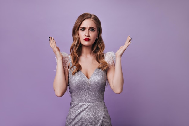 Woman in silver dress looking into camera with misunderstanding Sad lady in shiny outfit posing on isolated Portrait of blonde girl with red lips on purple backdrop