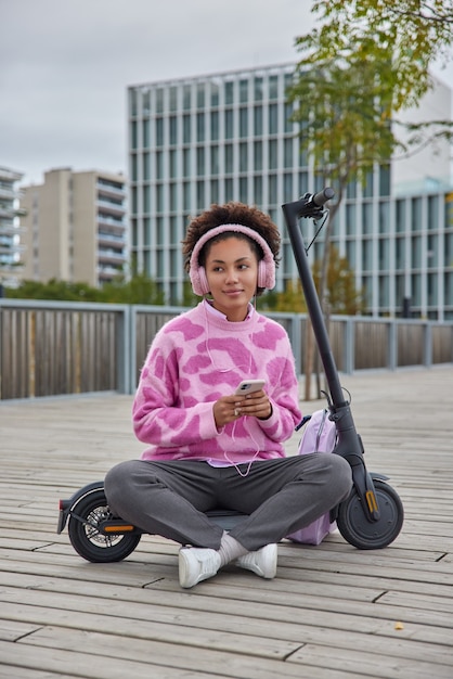 woman sits crossed legs listens favorite music in earphones wears casual jumper trousers and sneakers rides electric kick scooter poses at urban setting