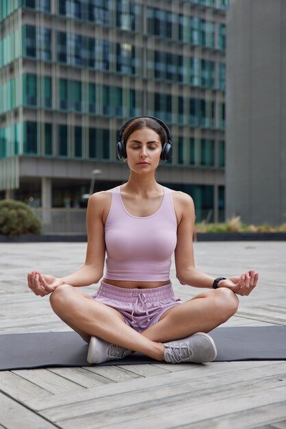女性はカレマットの練習で蓮華座に座るヨガは都市のスクレーパーの間でワイヤレスヘッドフォンで音楽を聴き、調和が深く呼吸するのを見つけます