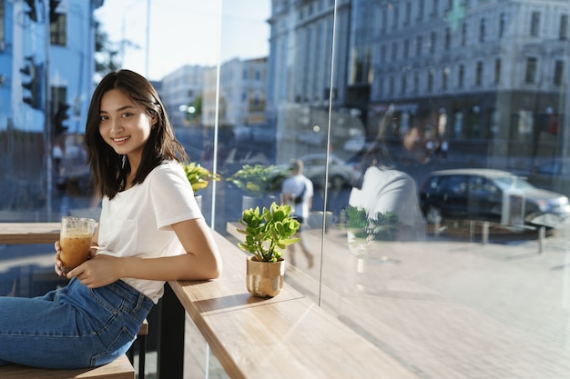 Free photo woman sits near counter and drink coffee, behind passersby and cars on streets, smiling joyfully