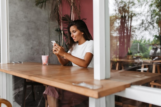 Woman sits at table in cozy cafe and chats in smartphone Portrait of tanned girl in white Tshirt