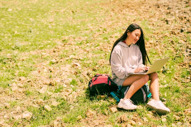 Woman sitting on backpack smiling and working on laptop in green hill