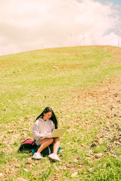 Foto gratuita donna che si siede sullo zaino e che lavora in computer portatile sulla collina verde