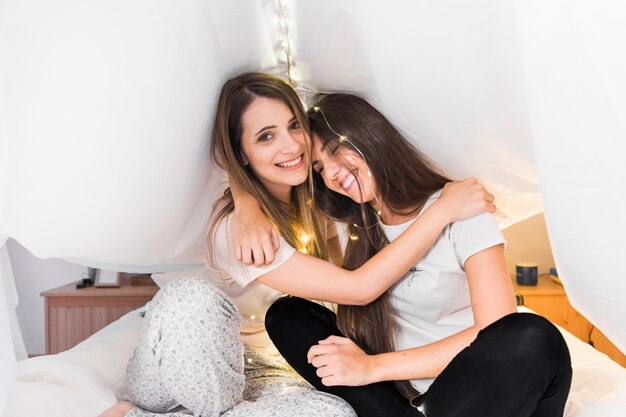 Woman sitting on bed hugging her female friend under the curtain