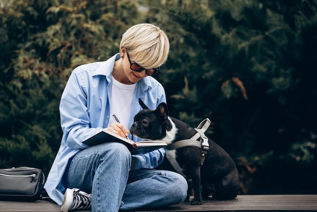 Woman sitting on a bench with french bulldog and writing in notebook