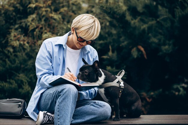 Woman sitting on a bench with french bulldog and writing in notebook