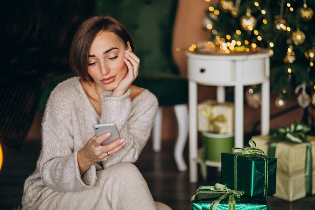 Woman sitting by Christmas tree and shopping on the phone