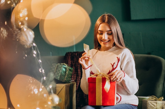 Woman sitting by Christmas tree and unpacking christmas present
