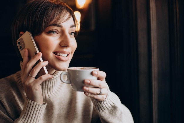 Женщина сидит в кафе, пьет кофе и разговаривает по телефону