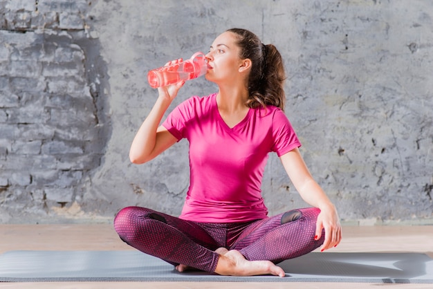 Woman sitting on exercise mat with crossed leg drinking water from bottle