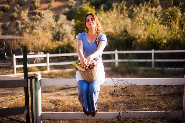 Woman sitting on the fence holding basket of harvested vegetable