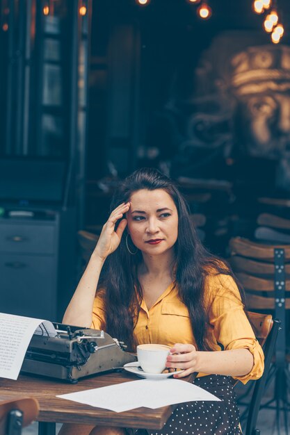 woman sitting and holding coffee in cafe terrace in yellow top and long skirt during daytime and looking thoughtful