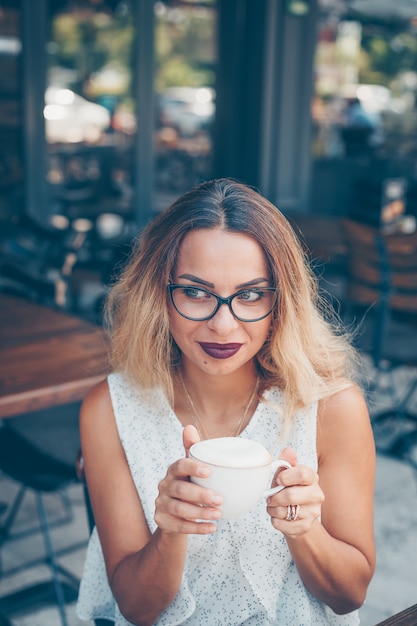 座っていると、日中のカフェのテラスでコーヒーと白い織り目加工シャツの女性。