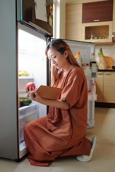 Женщина сидит перед открытым холодильником и проверяет, есть ли у нее все ингредиенты для блюда, которое она собирается приготовить