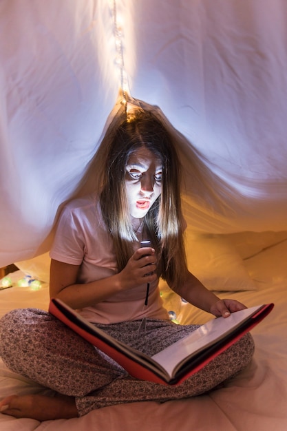 無料写真 彼女の顔の上にトーチの光を保持しているカーテンの読書雑誌の下でベッドの中に座っている女性