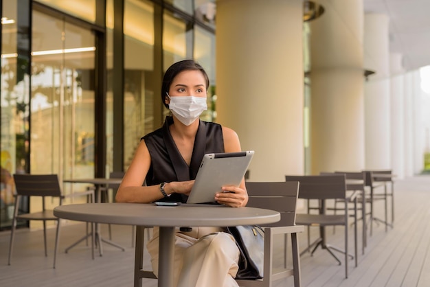 Женщина, сидящая на открытом воздухе в кафе-ресторане, социальное дистанцирование и ношение маски для защиты от covid 19 при использовании телефона и цифрового планшета горизонтального снимка.