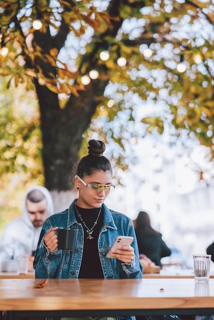 Woman sitting outdoors cafe drinking beverage holding in hand smartphone