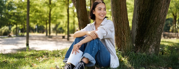 Free photo woman sitting in park with her favourite book leaning on tree under shade on sunny day enjoying