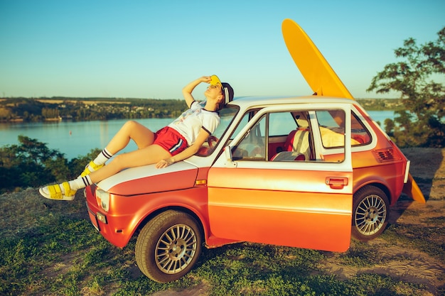 Woman sitting and resting on car on the beach on a summer day near river.