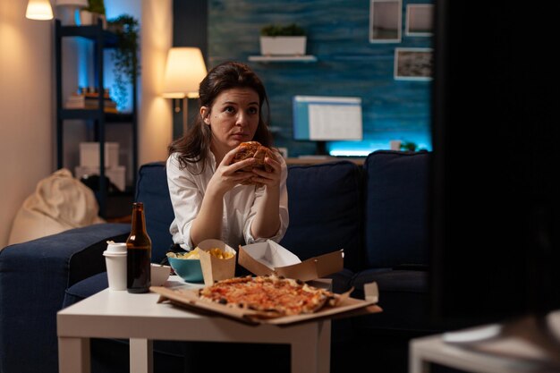 大きなピザとコーヒーテーブルの前でテレビでニュースを見ているおいしいおいしいハンバーガーを食べてソファに座っている女性。テイクアウトのジャンクフードの注文を楽しんでいる白人女性。