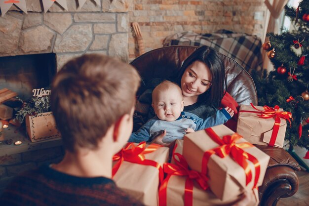 Женщина, сидящая на диване с ребенком на руках и с горой подарков коричневых с красным бантом