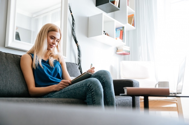 Foto gratuita donna che si siede sul sofà e che scrive in blocco note a casa