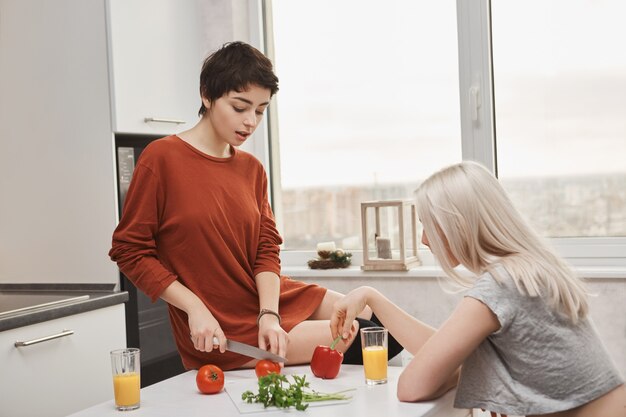 Woman sitting on table cutting tomotoes while her friend drinks orange juice