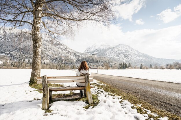 木製のベンチに座って、曇り空の下で雪に覆われた山々を見ている女性