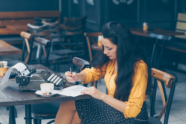 Free photo woman sitting and writing something on paper in cafe terrace in yellow top and long skirt during daytime and looking thoughtful
