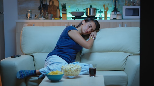 Женщина спит на диване перед телевизором во время просмотра скучающего фильма. Усталая, измученная одинокая сонная дама в пижаме засыпает, сидя на уютном диване в гостиной, закрыв глаза по ночам