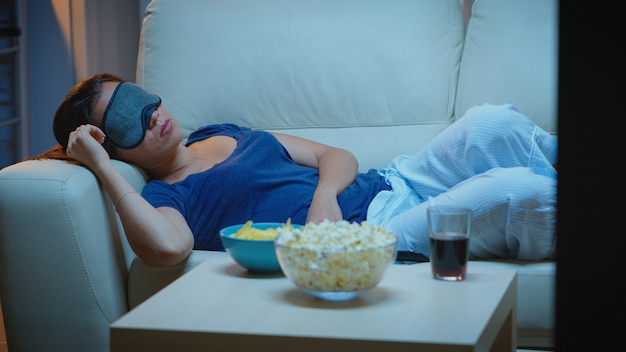 Женщина спит с маской покрытия глаз перед телевизором, лежа на диване. Усталая, измученная одинокая сонная дама в пижаме засыпает на диване перед телевизором, закрыв глаза во время просмотра фильма.