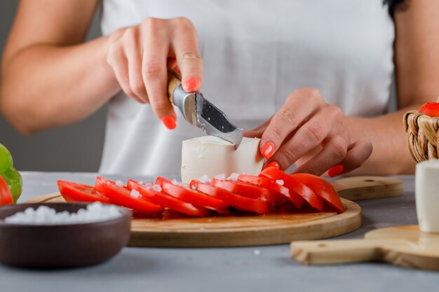 Woman slicing cheese in cutting board with sliced tomatoes, salt on gray surface