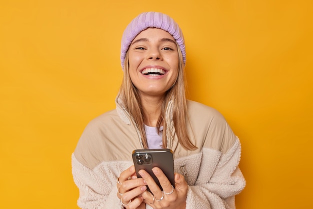 woman smiles broadly enjoys modern technologies uses smartphone for online communication wears knitted hat and jacket isolated on yellow downloads cool app