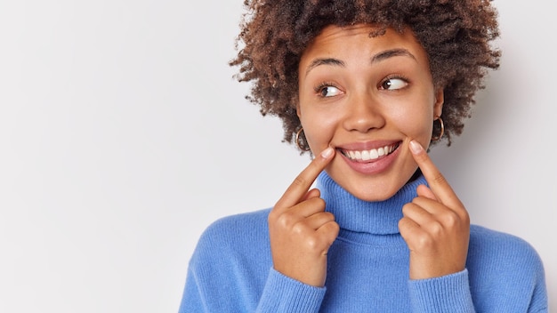 woman smiles broadly points at her perfect even teeth indicates at corners of lips looks away wears casual blue jumper isolated on white with blank copy space.