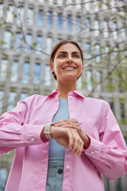 женщина нежно улыбается, проверяет время на наручных часах, ожидает, когда кто-то придет на встречу, в розовой рубашке позирует на фоне городского здания
