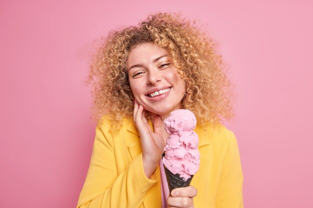 女性の笑顔が優しく頭を傾ける明るい夏の日の暑い夏の日に、おしゃれな黄色のジャケットのポーズを着て屋内でおいしいピンクのアイスクリームを食べます。おいしいデザート