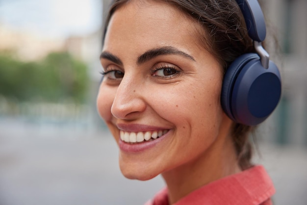 Woman smiles toothily enjoys leisure time listens favorite music in wireless headphones poses outdoors outdoor