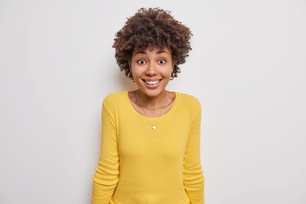 女性は歯を見せて微笑んで、驚いたように見えます。素晴らしいニュースが白にカジュアルな黄色のセーターのポーズを着ているのを聞きます。人間の顔の表情感情と感情の概念