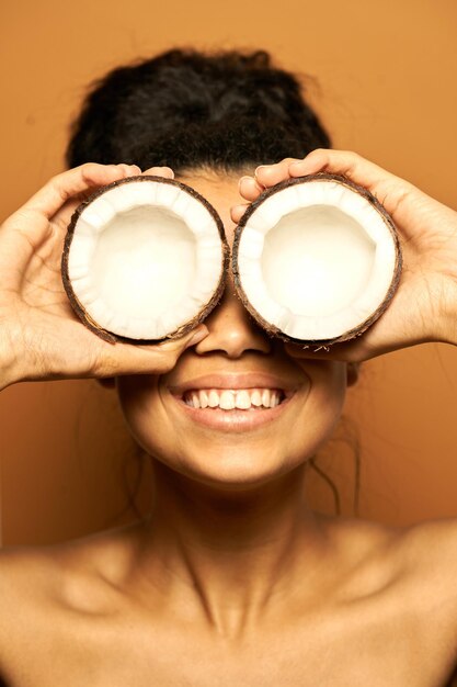 woman smiling at camera, holding two coconut halves on her eyes, posing isolated on orange