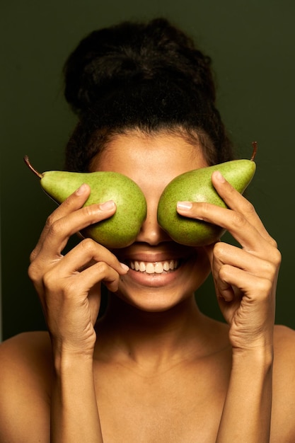woman smiling at camera, holding two juicy pears on her eyes, posing isolated on green