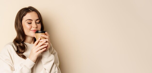 Woman smiling and smelling delicious cup of coffee in takeaway cup standing over beige background