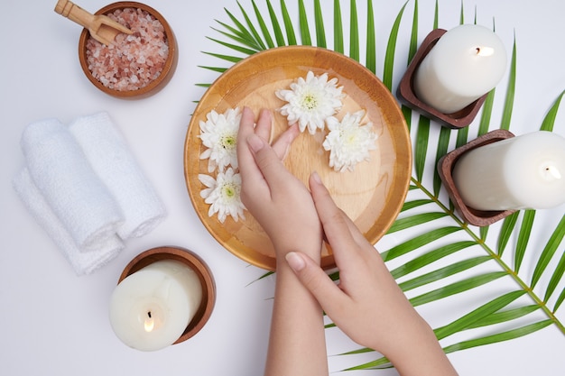 Donna che immerge le mani in una ciotola di acqua e fiori, trattamento spa e prodotto per piedi femminili e spa per le mani, massaggio con ciottoli, acqua profumata di fiori e candele, relax. lay piatto. vista dall'alto.