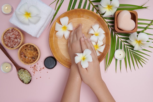 Woman soaking her hands in bowl of water and flowers, Spa treatment and product for female feet and hand spa, massage pebble, perfumed flowers water and candles, Relaxation. Flat lay. top view.