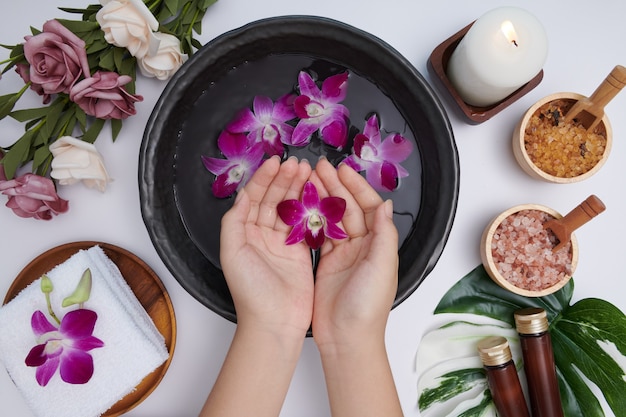 Woman soaking her hands in bowl of water and flowers, Spa treatment and product for female feet and hand spa, massage pebble, perfumed flowers water and candles, Relaxation. Flat lay. top view.