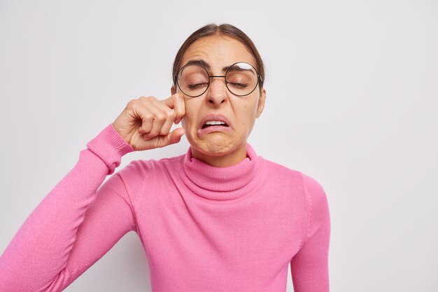 woman sobs and wipes tears being very upset about something wears transparent glasses poloneck expresses negative emotions on white cries from hopelessness