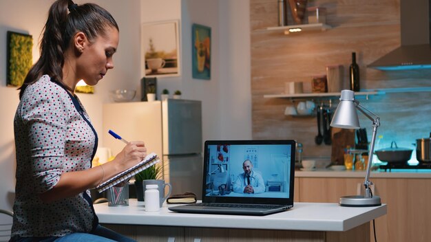 Woman speaking online with doctor taking notes during tele health sitting in home kitchen. Sick lady discussing during virtual consultation about symptoms holding notebook and writing treatment