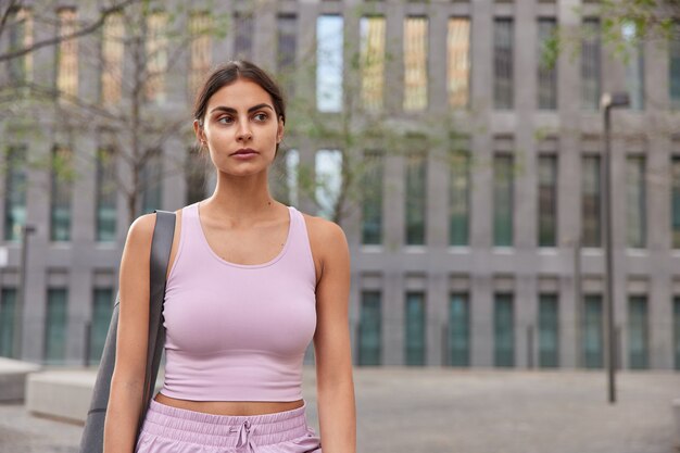  woman in sportswear focused into distance carries rubber rolled karemat poses on modern building prepares for workout session