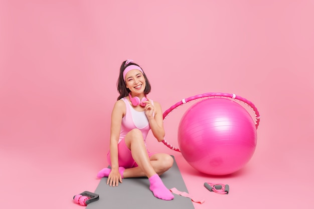 woman in sportswear poses on fitness mat wears headphones around neck has sincere glad expression trains with swiss ball and hula hoop isolated on pink 