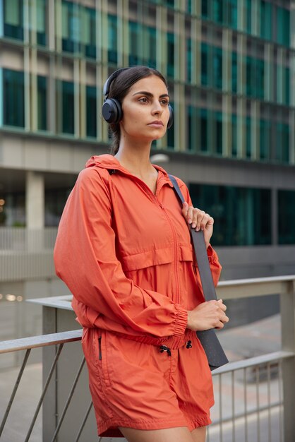  woman in sportswear waits for coach outdoors has break after workout or jogging exercises every day to be healthy rests and looks forward enjoys favorite playlist